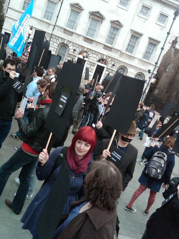 A woman with dyed red hair holds up a placard. It is completely black. In the background, protestors hold up similarly black signs.